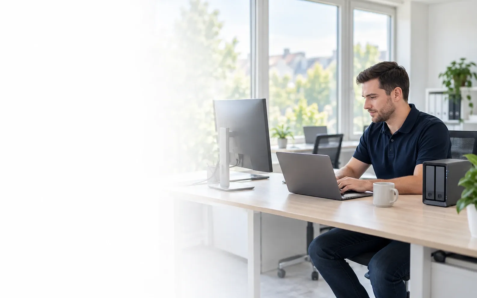 IT-Betreuer arbeitet am Laptop in einem hellen Büro, daneben Monitor und NAS-Speicher